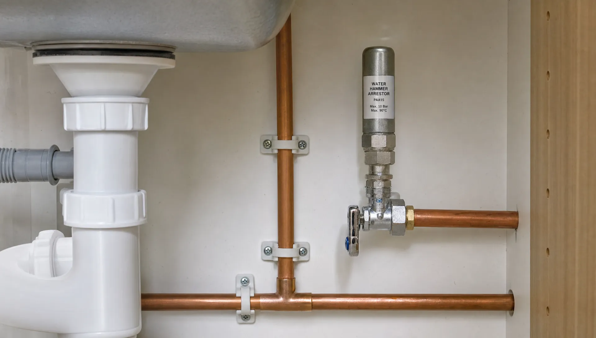 A cutaway view of household water pipes under a kitchen sink, showing a small water hammer arrestor fitted near an appliance valve and secure pipe clips holding the pipework in place.