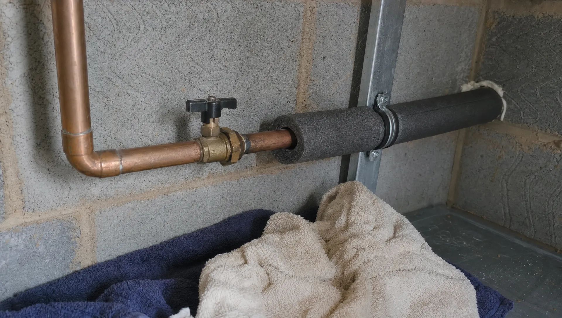 Exposed pipework in a cold utility space shows foam lagging partly fitted, a nearby stopcock and towels ready below to catch a leak.